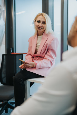 Confident woman in pink blazer smiles while using smartphone in a modern office settingの写真素材