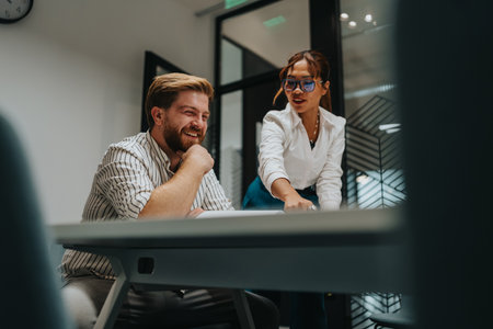 Two colleagues collaborate at a modern office desk, discussing work and sharing a moment of laughter