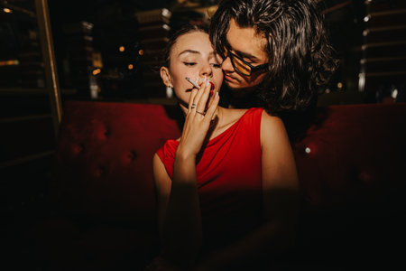 Intimate night out with friends: woman in red dress smokes while friend leans closeの写真素材