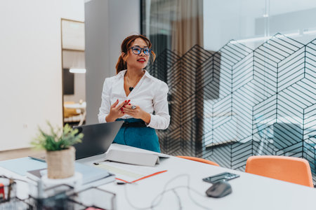 Confident multicultural businesswoman presenting in a modern office with laptop, glass partition, and organized deskの写真素材