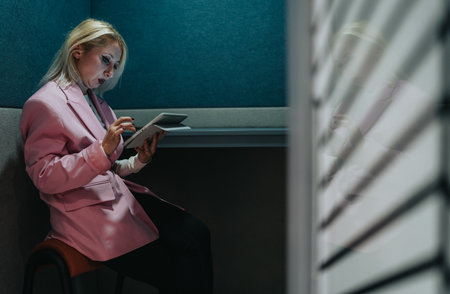 Businesswoman in a pink blazer using tablet in a quiet booth for focused workの写真素材
