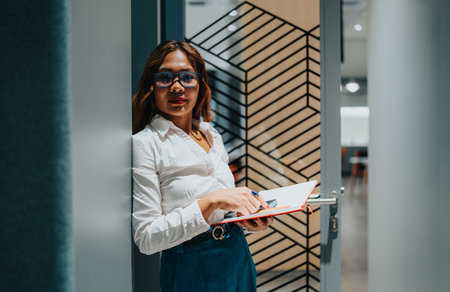 Confident woman in a white shirt holds a notebook at the office door, ready to plan and work