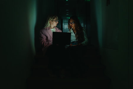 Two women collaborate on a laptop in a dim hallway stairwell at night