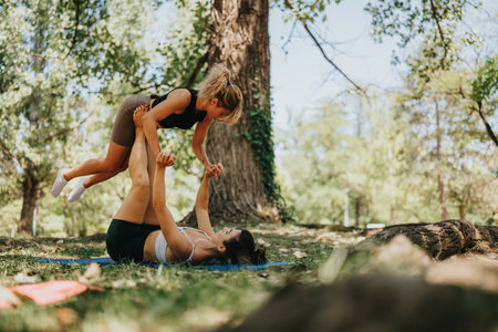 Two women perform partner acro yoga outdoors in a sunny park, balancing and supporting each other.