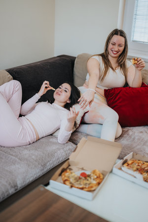 Two young women enjoying pizza together while lounging on a cozy sofaの写真素材
