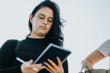 Young woman outdoors writing in a notebook with pen and glasses, focused and thoughtfulの写真素材