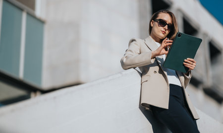 Professional woman in beige blazer using tablet outdoors on a sunny city dayの写真素材