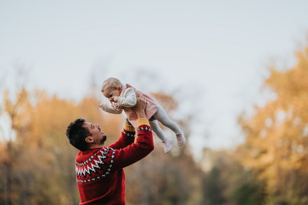 Man in red sweater lifts baby in autumn park, a joyful outdoor family momentの写真素材