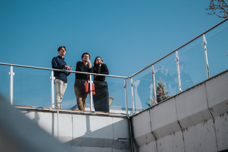 Three young professionals stand on a glass railing outdoors, overlooking a sunny blue sky and modern railingの写真素材