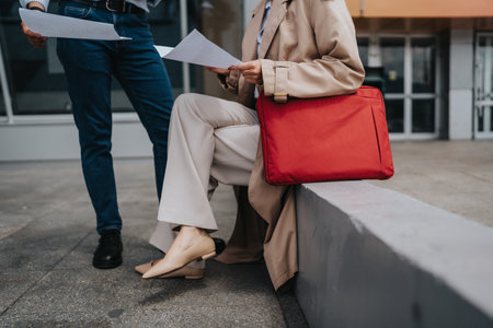 Business colleagues reviewing documents outdoors with red bag on a concrete seatの写真素材