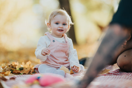 Baby girl in pink overalls sits on a blanket in autumn park with family nearby