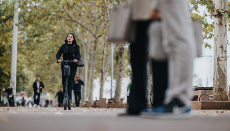 Woman in black coat rides electric scooter along tree-lined city street while pedestrians pass byの写真素材