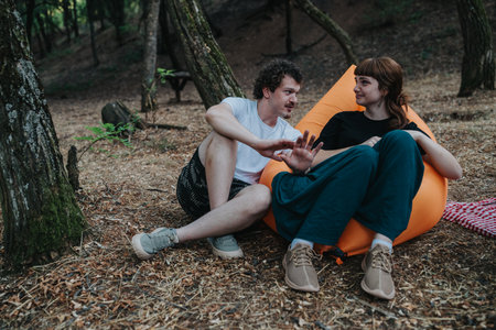 Friends relax in a forest campsite on an orange beanbag chair, sharing a casual conversationの写真素材