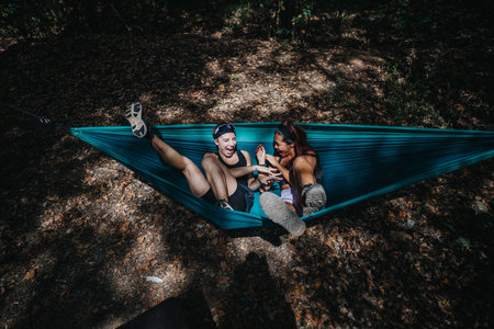 Two friends laugh and relax in a blue hammock hanging between trees in the forestの写真素材