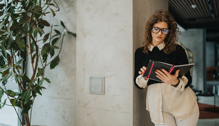 Professional woman in office holds notebook and tablet while leaning against wall during a workdayの写真素材