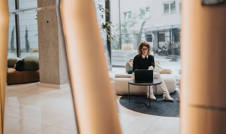 Woman working on laptop in a modern coworking lounge with comfy seating and large windowsの写真素材