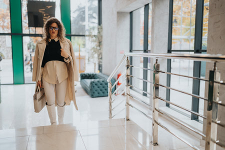 Professional woman in beige trench coat ascending stairs in a modern office lobbyの写真素材