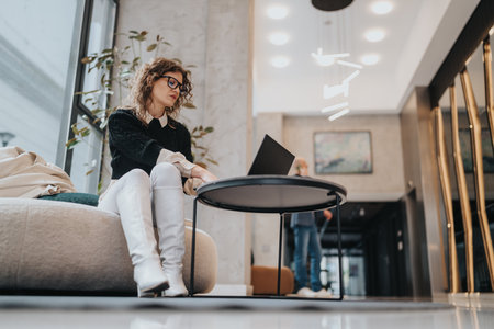 Woman working on laptop in a modern lobby lounge with stylish office decor and calm business vibeの写真素材