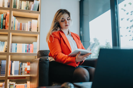 Businesswoman in an orange blazer reads a book beside a laptop in a cozy bookstoreの写真素材