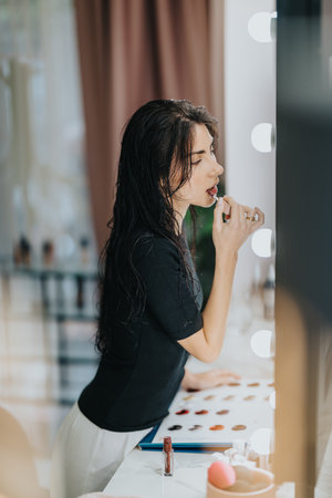 Woman applying lipstick at vanity mirror with makeup palette and bright lightsの写真素材