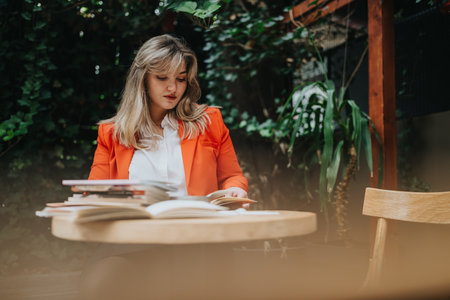 Woman in orange blazer reads books at an outdoor cafe, surrounded by greenery and calm ambianceの写真素材