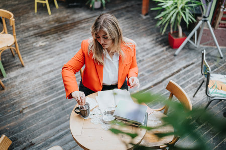 Businesswoman in orange blazer working at an outdoor cafe with papers and coffeeの写真素材