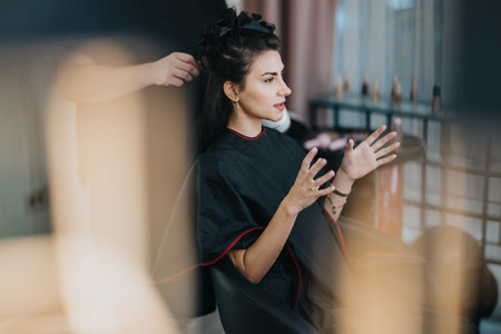 Woman in a hair salon receiving a styling session with cape on and expressive hand gesturesの写真素材