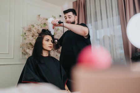 Woman sits in a salon chair as stylist curls her hair with a curling iron for a makeoverの写真素材