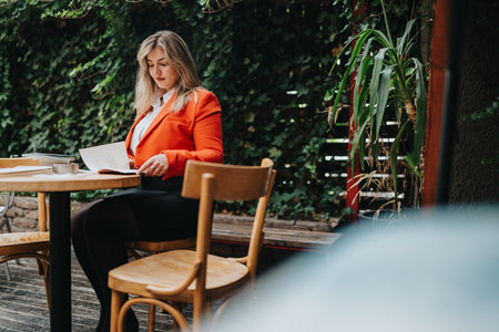 Woman in red blazer reads papers at outdoor cafe surrounded by greeneryの写真素材