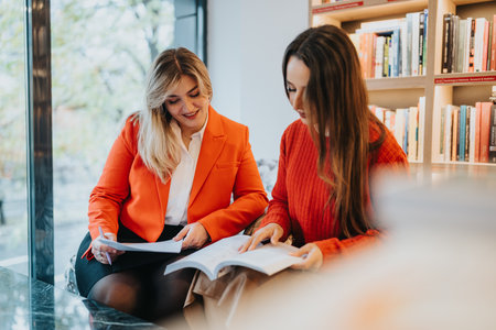 Two women study together in a bookstore, sharing ideas and examining books by the windowの写真素材