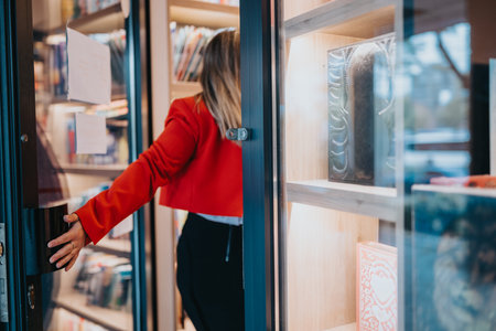 Woman in red jacket enters a bookstore, reaching for the door as shelves fill with booksの写真素材