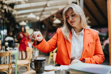 Stylish woman in an orange blazer works at a cafe desk with coffee and documentsの写真素材