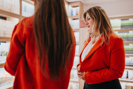 Two women in red jackets discuss books in a bright bookstore setting during a shopping tripの写真素材