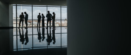 Silhouetted colleagues gather in a bright glass-walled office lobby for a casual business meetingの写真素材