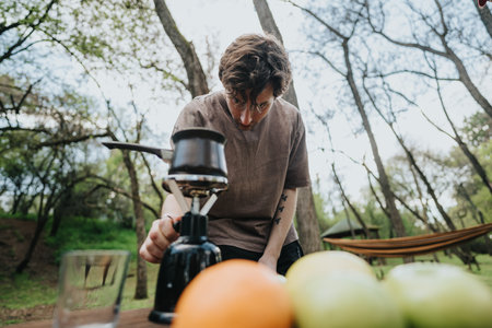 Young man camping outdoors, cooking with a portable stove among trees and a hammock in natureの写真素材