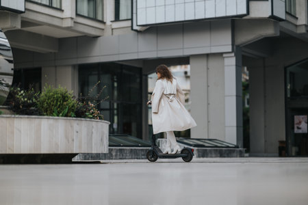 Woman in beige trench coat rides an electric scooter through a modern urban city sceneの写真素材