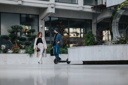 Stylish couple walking outdoors in a modern urban plaza with a man riding a scooterの写真素材