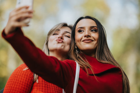 Two friends take a cheerful selfie outdoors in fall colors, wearing red clothing and smilesの写真素材