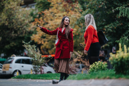 Two women in autumn outfits chat on a city park path, wearing red coats and stylish outfitsの写真素材