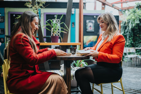 Two women in red jackets share a conversation over books at a colorful cafe outdoorの写真素材