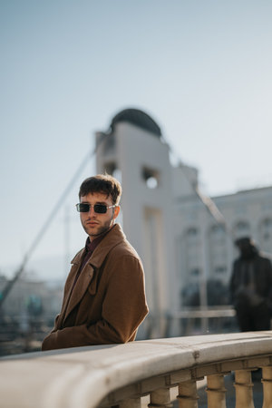 Young professional man on a city bridge in a brown coat wearing sunglasses, exuding confidence and styleの写真素材