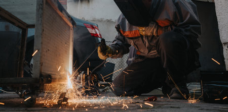 Welding worker kneeling and cutting metal, sparks flying in a gritty industrial workshop sceneの写真素材