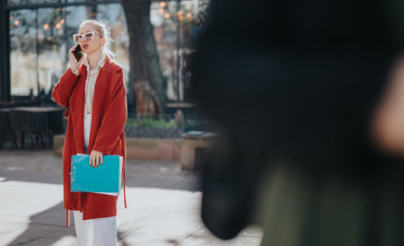 Confident businesswoman in red coat speaks on the phone while holding a blue clipboard outdoorsの写真素材