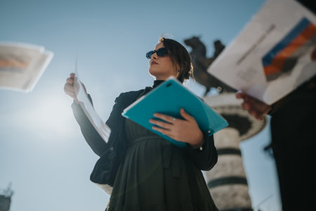 Confident businesswoman in urban setting presents documents and tablet under bright sunの写真素材