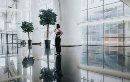 Business woman stands in a bright modern atrium with polished floor reflections and greeneryの写真素材