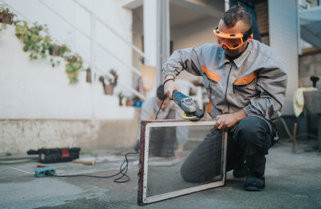 Focused worker using power tool to sand a glass frame outdoors with safety gogglesの写真素材