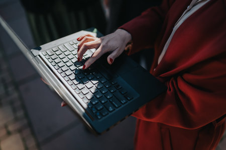 Businessperson typing on a laptop outdoors in the city with a red coat and focused handsの写真素材