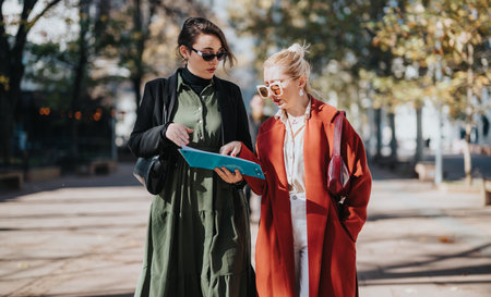 Two stylish women in a city street share a clipboard and discuss fashion and businessの写真素材