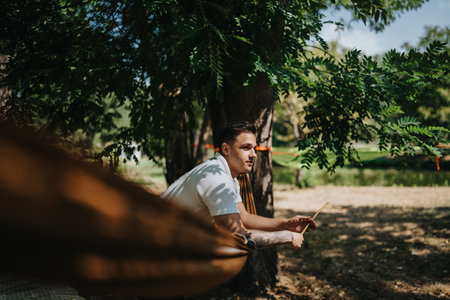 Relaxed man lounging in a hammock under a tree in a sunny park settingの写真素材