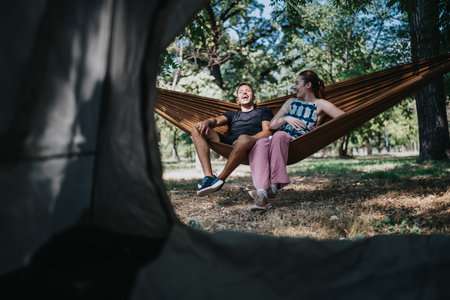 Friends laughing in a hammock between trees during a sunny camping trip, enjoying outdoor relaxationの写真素材